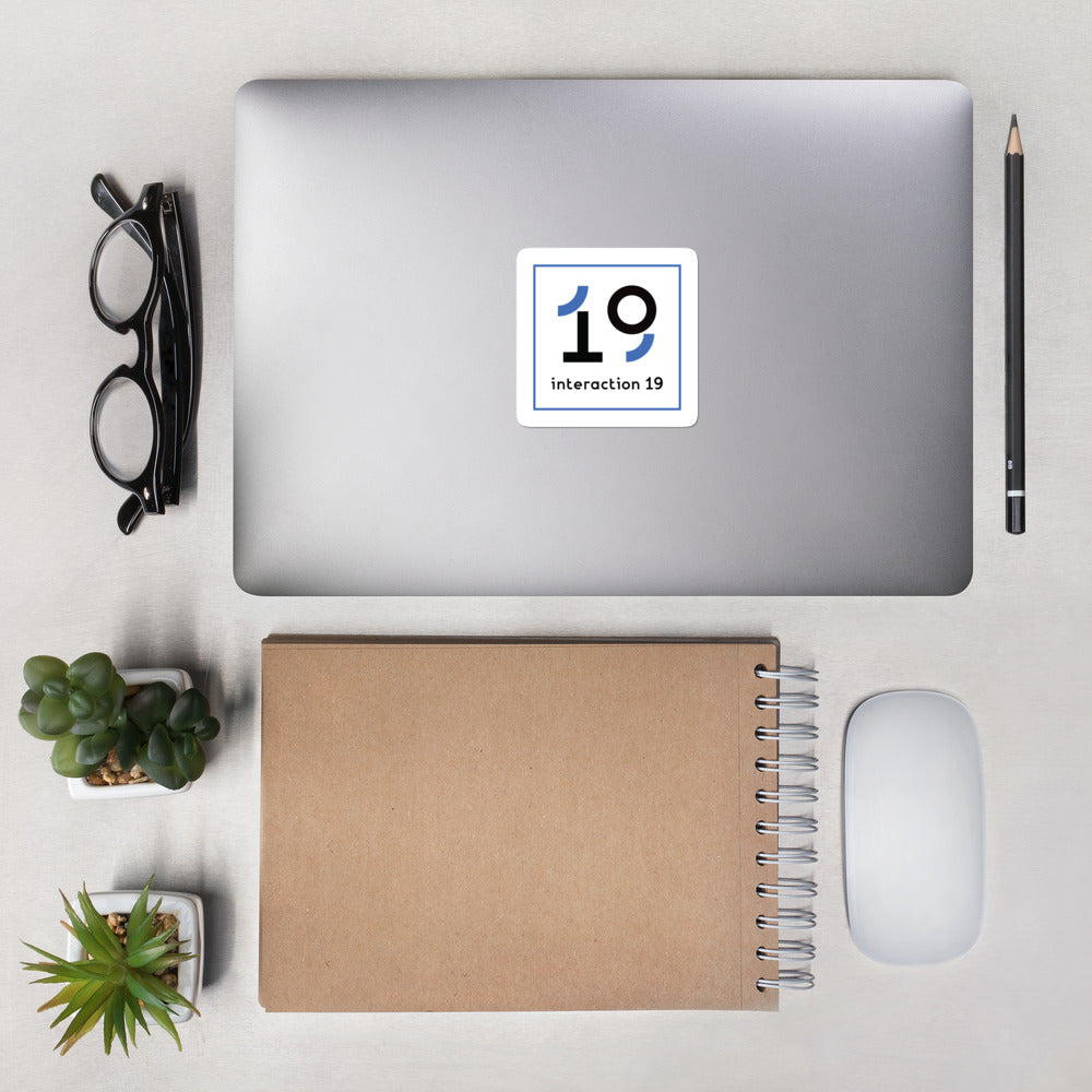 Square white sticker with blue and black Interaction 19 logo on surface of a laptop shot from above. Glasses, pencil, notebook, two small plants and computer mouse are also on table top.