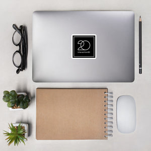 Square black sticker with white Interaction 20 logo on surface of a laptop shot from above. Glasses, pencil, notebook, two small plants and computer mouse are also on table top.